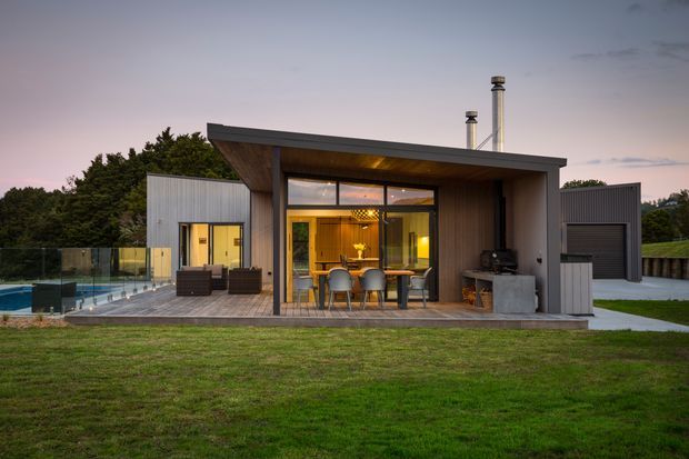 A contemporary single-storey pavilion-style home viewed from the lawn at dusk. The building has a broad asymmetric flat roof with deep overhangs, dark vertical timber cladding, corrugated metal side volumes, extensive floor-to-ceiling glazing, and a covered timber deck furnished with an outdoor dining table and chairs. A swimming pool with glass balustrade is visible to the left, along with chimney flues and landscaped grounds.