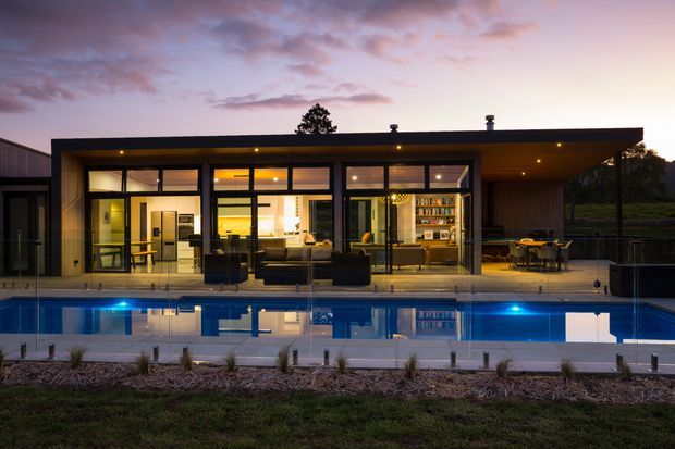 A low-slung, single-storey modern house viewed from across a rectangular swimming pool at dusk. The facade features extensive floor-to-ceiling glazing in dark frames, warm timber soffit lining, a flat roof, and an open terrace with outdoor dining furniture. Through the glazing, a warmly lit open-plan living and kitchen area is visible. The pool dominates the foreground and reflects the illuminated house and blue accent lighting.