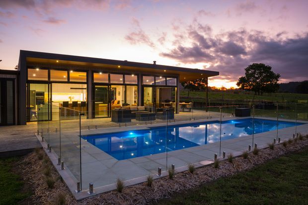 A contemporary single-storey pavilion-style home viewed at dusk, with extensive floor-to-ceiling glazing opening onto a tiled terrace and rectangular swimming pool. The dark low-pitched roof, slim dark window frames, illuminated open-plan interior, glass pool fencing, and landscaped rural grounds create a refined indoor-outdoor setting.