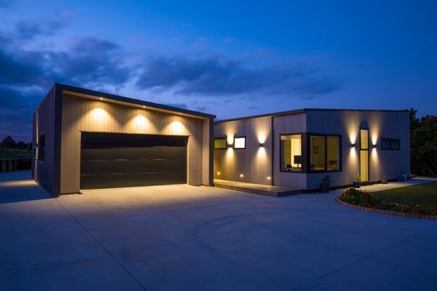 A modern single-storey residential property photographed at dusk, with a large detached-looking garage volume connected to a low rectangular home. The facade combines dark vertical cladding with warm timber-toned vertical panels, broad minimal glazing, and multiple exterior wall lights. A wide concrete driveway and paved forecourt dominate the foreground, with a small landscaped garden beside the entrance.