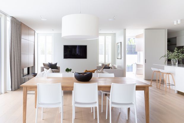 Bright open-plan contemporary interior featuring a timber dining table with white chairs, a white kitchen island with timber bar stools, and a living area beyond. The space has light timber flooring, white walls and cabinetry, integrated timber wall joinery, large windows with sheer curtains and plantation shutters, and a prominent oversized white pendant light.