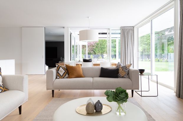 Bright, contemporary living room with pale timber flooring, a light upholstered sofa, patterned and ochre cushions, a round white coffee table, and extensive floor-to-ceiling glazing overlooking a garden. An open dining area and kitchen are visible beyond the seating zone, with neutral curtains and a large pendant light.
