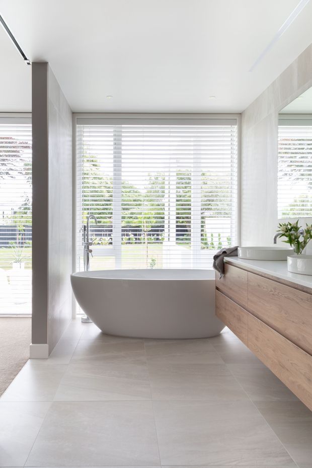 Bright contemporary bathroom featuring a freestanding white bathtub beneath a large window with white horizontal blinds, a timber floating vanity with an integrated white basin, pale large-format floor and wall tiles, and a large wall mirror. Natural greenery is visible through the glazing.