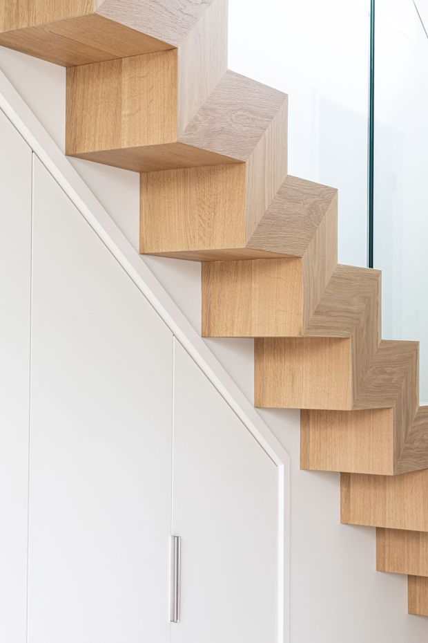 Close-up architectural detail of a sculptural, open-riser staircase formed from chunky light timber treads and stepped side elements, set against a clean white wall. A white built-in storage door with a slim metal handle sits beneath the stair, while a vertical glass balustrade or screen is visible at the right edge. The composition is bright, minimalist, and highly geometric.