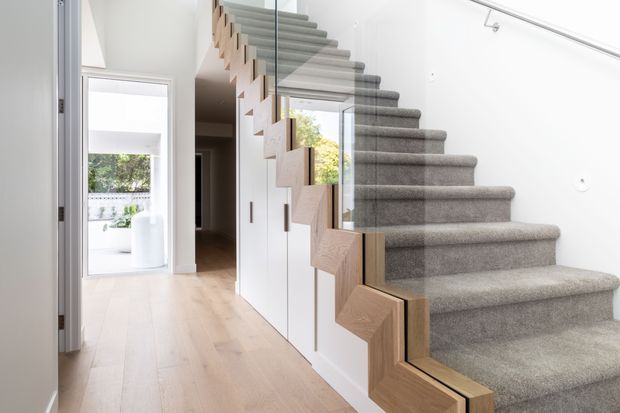 Bright contemporary hallway featuring a floating or open-riser staircase with grey carpeted treads, pale timber zigzag stringers, and a clear glass balustrade. White walls and cabinetry line the stair, while a light timber floor leads toward a bright adjoining space.