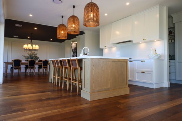 Spacious open-plan kitchen and dining area with white shaker-style cabinetry, a large timber-panelled island with bar seating, dark timber flooring, and woven pendant lights. A formal dining table is visible behind the kitchen, beneath a dark ceiling feature with recessed lighting. The room has a polished, warm, classic residential interior.