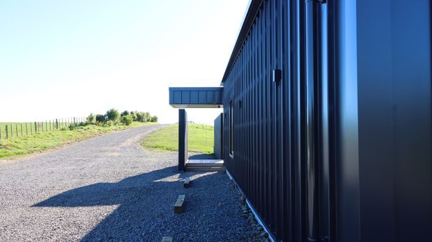 Side view of a contemporary dark-clad building in a rural setting, with a long corrugated metal wall, a projecting covered entry or canopy, gravel driveway, and open grassy fields beyond. Strong linear perspective and crisp daylight emphasize the building’s simple industrial form.