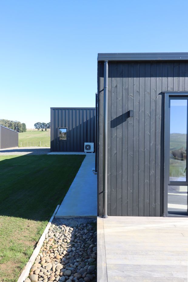 Exterior view of a modern low-rise building composed of dark vertical timber cladding with simple black metal trim and a large glazed door or window. A pale timber deck, concrete walkway, gravel drainage strip, and manicured lawn surround the building. Additional dark-clad structures and open rural farmland are visible in the background under a clear blue sky.