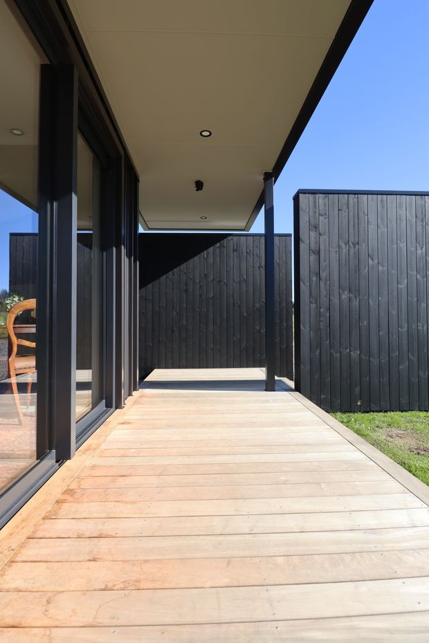A minimalist covered outdoor deck or terrace beside a glazed modern building. The deck is finished in pale timber boards, with dark-framed floor-to-ceiling glazing on one side and tall dark vertical timber privacy screens enclosing the space. A flat soffit with recessed downlights extends overhead, creating a sheltered walkway or outdoor living area.