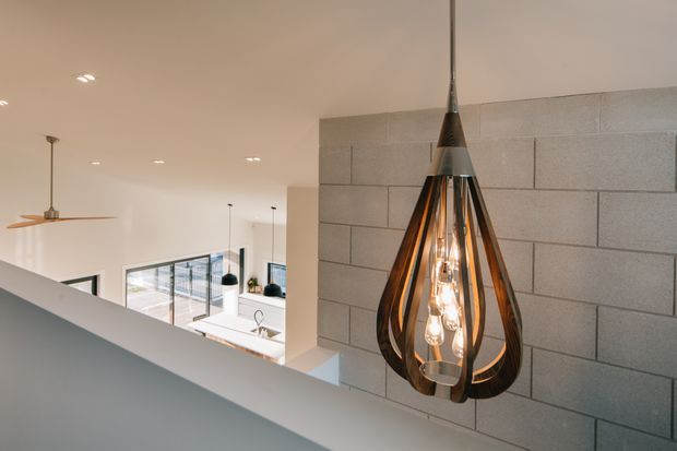 Interior view of a contemporary multi-level home, focused on a sculptural teardrop pendant light hanging in front of a grey concrete block feature wall. A bright open-plan kitchen and living area is visible beyond the stair or landing edge, with large windows, recessed ceiling lights, and a ceiling fan.