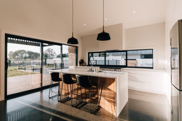 Bright contemporary kitchen with a large white island, timber feature cabinetry, integrated cooktop and sink, three black bar stools, pendant lights, and expansive black-framed glazing opening to an outdoor deck. Polished dark flooring and warm neutral walls create a refined, inviting interior.
