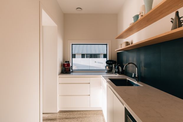 A compact contemporary galley kitchen with white flat-panel cabinetry, a pale stone benchtop, a dark teal backsplash, open timber shelving, and integrated appliances. A window above the sink provides natural light and overlooks a nearby corrugated-metal roof.
