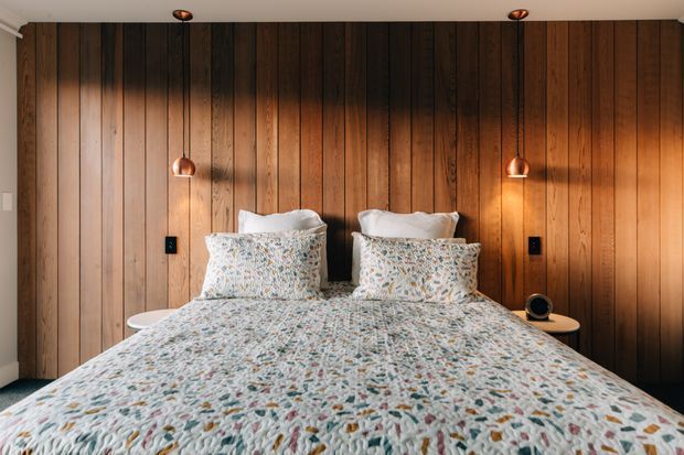 A warm, cozy bedroom centered on a neatly made bed with a floral-patterned quilt and matching pillows. The bed is set against a full-height dark timber vertical-panel feature wall, with integrated black switches, small rounded bedside tables, and symmetrical copper-toned pendant lights. Recessed ceiling spotlights provide additional illumination.
