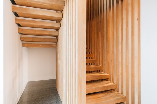 A bright interior hallway with a timber staircase. The stair is formed from open wooden treads and chunky exposed beams, enclosed by vertical timber slat screening. A carpeted passage runs alongside the stair, with pale walls and warm natural wood throughout.