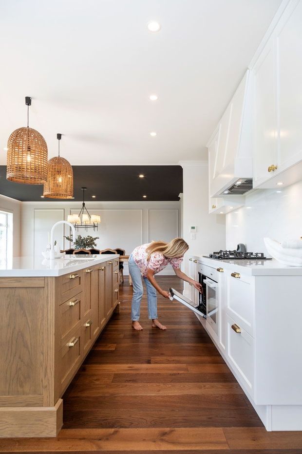 Bright, spacious kitchen with white shaker-style cabinetry, brass hardware, a dark stone or composite island benchtop, and a large timber island base. The room has dark timber flooring, white panelled walls, recessed ceiling lights, woven pendant lights, and a black ceiling feature over the dining area. A gas cooktop, oven, rangehood, sink, and dining space are visible, creating a warm contemporary-traditional kitchen interior.