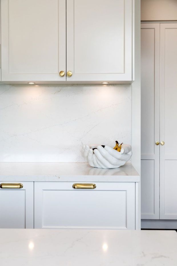 Close-up of a refined white kitchen with Shaker-style framed cabinetry, brass knobs and pulls, integrated under-cabinet lighting, and pale veined stone surfaces. A decorative bowl sits on the counter, while tall pantry-style cabinetry is visible at right. The image has a bright, clean, softly traditional look with subtle gold accents.