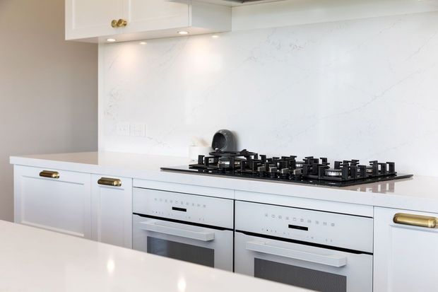 Bright, refined contemporary kitchen featuring white shaker-style cabinetry with brass pulls, a large white veined stone splashback and benchtops, a wide black gas cooktop, and twin built-in white ovens. Under-cabinet lighting and a glossy foreground island create a clean, polished appearance.