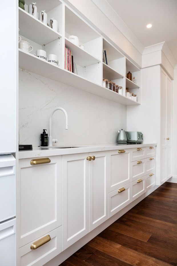 Bright, refined kitchen or butler's pantry with a long run of white shaker-style cabinetry, brass pulls, a white stone benchtop and matching veined splashback. Open white shelving displays ceramics, books and kitchenware, while a white gooseneck tap, sink and muted green kettle and toaster sit along the counter. Dark timber flooring and decorative wall moulding add warmth and classic detail.