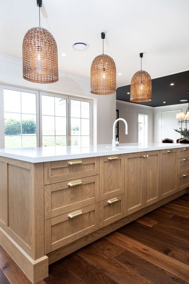 Bright kitchen interior centered on a large island with detailed oak cabinetry, brass hardware, and a white benchtop with an integrated sink and curved white tap. Three woven pendant lights hang above the island. Dark timber flooring, white walls and ceiling, large glazed doors, and a dark ceiling section create a warm, refined contemporary look.