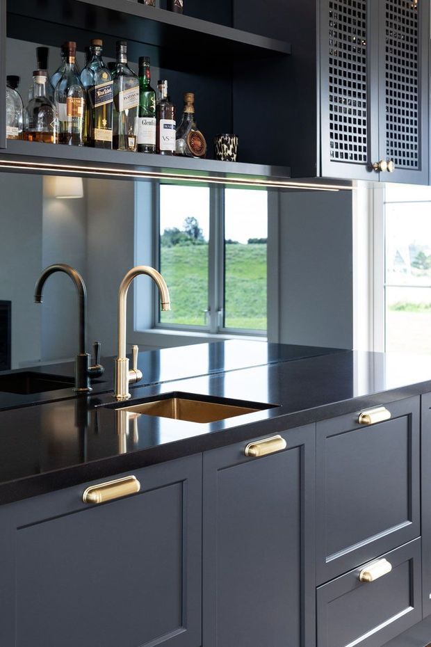 A refined dark-toned kitchen and home bar area with matte charcoal-blue shaker-style cabinetry, polished dark stone benchtops, brass pulls, and two tall arched faucets over an undermount sink. Open shelving displays liquor bottles, while a bright window looks onto a green rural landscape. A decorative perforated cabinet door and integrated under-shelf lighting add detail.
