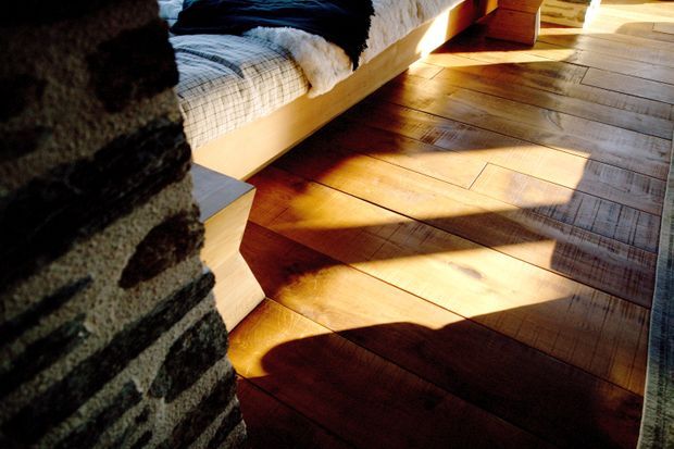 Close-up interior view of a bedroom, focusing on warm timber plank flooring illuminated by strong sunlight and shadows. A low timber bed with layered bedding is visible at the top, alongside a dark textured masonry or stone feature wall at left and a small timber bedside surface. The space has a rustic, tactile and warmly lit character.