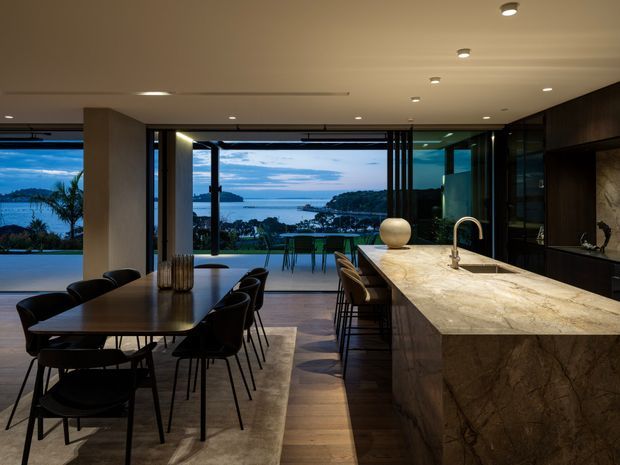 A sophisticated open-plan kitchen and dining area with expansive glazing overlooking a coastal bay. A large stone island with an integrated sink and curved brass-toned tap dominates the right side, while a long dark dining table with black chairs occupies the left. Dark timber cabinetry, warm recessed lighting, timber flooring, and a broad neutral rug create a refined, moody interior with strong indoor-outdoor connection.