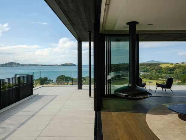 A contemporary open-plan living area opening onto a broad tiled terrace with black metal balustrades and expansive floor-to-ceiling glazing. The interior features timber flooring, a sculptural suspended black fireplace, sparse lounge furniture, and strong visual connections to a coastal inlet and rolling landscape beyond.