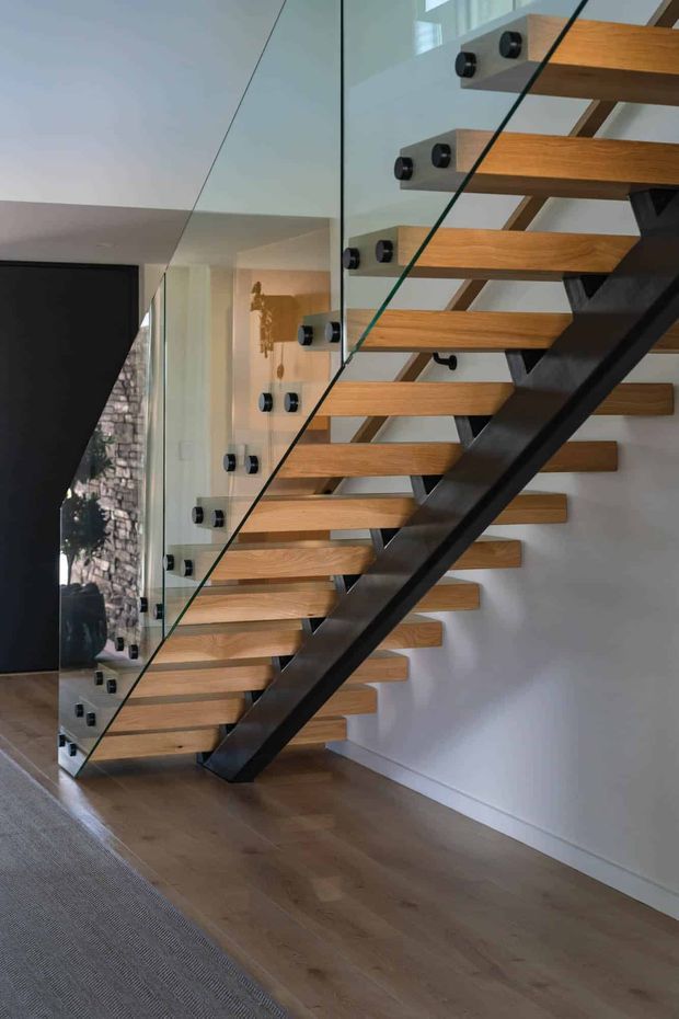 Interior view of a contemporary floating staircase with thick warm timber treads, a dark metal central stringer, and frameless glass balustrades secured by round metal fittings. The stair rises beside a white wall with matching timber wall-mounted tread ends, while a large dark opening and reflective glazing are visible to the side. The space has timber flooring and a clean, minimalist architectural character.