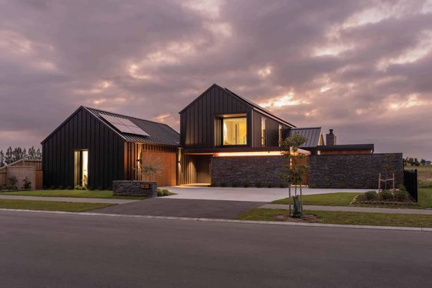 Street-facing dusk photograph of a contemporary multi-volume residential house. The home has steep gabled roof forms, dark vertical cladding, warm-lit windows and entry areas, a low stone-clad garage or boundary wall, a paved driveway, and a neatly landscaped suburban frontage under a dramatic cloudy sky.