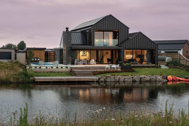 Exterior view of a contemporary dark-clad multi-gabled house beside a landscaped waterfront canal or pond. The home features extensive glazing, a prominent stone chimney, covered outdoor living areas, a timber deck and steps leading to a small private dock. Kayaks are stored at the water's edge, and warm interior lighting is visible through the windows.