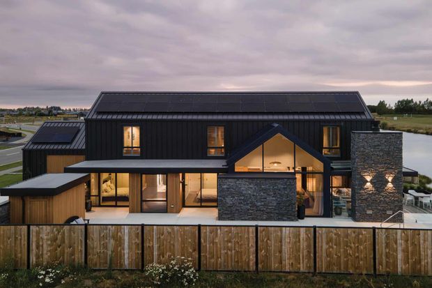 Exterior view of a contemporary, dark-clad multi-level house at dusk. The home features a long pitched roof, black vertical cladding, extensive glazing, a projecting gabled entry or living volume, and prominent stacked-stone chimneys or feature walls. A timber privacy fence encloses the foreground, with open rural or suburban landscape beyond.