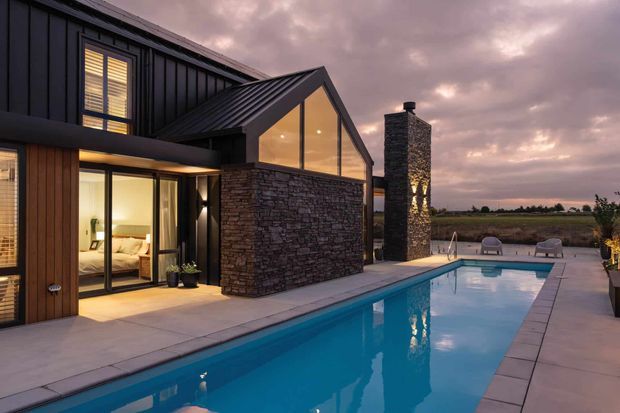A contemporary dark-clad house at dusk with a steep gabled glazed section, extensive natural stone walls, timber detailing, and a tall stone chimney. Floor-to-ceiling glazing reveals a warmly lit bedroom or living area. A long rectangular swimming pool and paved terrace occupy the foreground, with open rural fields beyond.