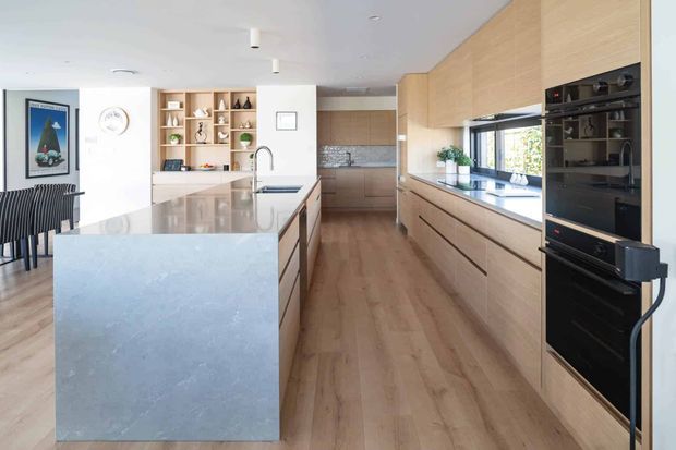 Bright contemporary kitchen with a large pale stone island, extensive light timber cabinetry, integrated black appliances, open shelving, and a dining area visible at the left. The room has a long linear layout, timber flooring, white walls and ceiling, and generous natural light through the window above the benchtop.