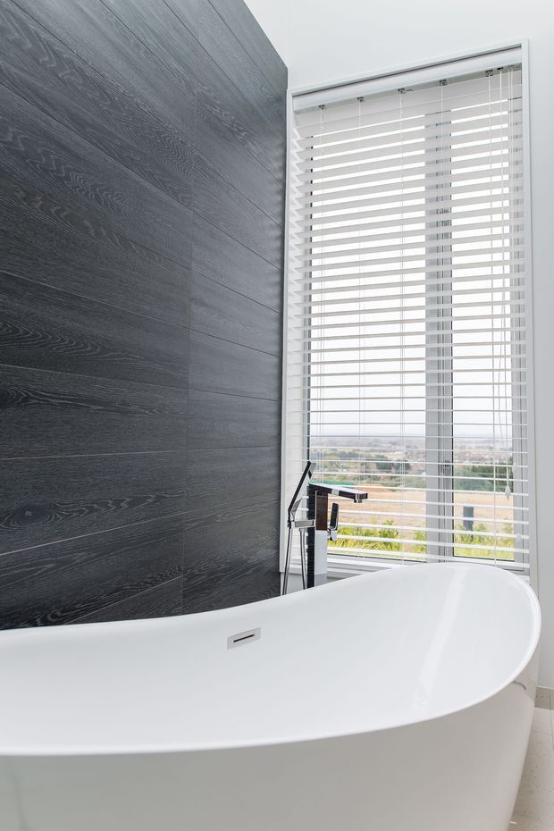 A contemporary bathroom featuring a freestanding white bathtub beside a tall window with white horizontal blinds. The wall behind the tub is clad in dark grey wood-look panels, with a minimalist chrome and black floor-mounted bath tap. A bright suburban or urban view is visible through the window.