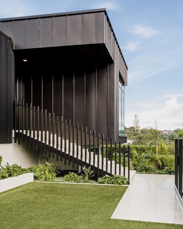 Exterior view of a contemporary multi-level building with a dark metal-clad upper volume projecting over a recessed entry or stair zone. Slim vertical metal balustrades line the sloping access path, with large glazed panels at the side. A pale concrete walkway, manicured lawn, and dense tropical planting frame the structure, with a suburban landscape visible beyond.