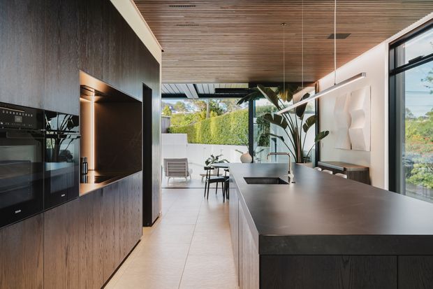 Open-plan contemporary kitchen with a large dark island, integrated black appliances, and full-height dark timber cabinetry. A warm timber slatted ceiling, pale tiled flooring, expansive glazing, and indoor tropical planting create a refined connection to the outdoor garden. A dining and lounge area is visible beyond the kitchen.