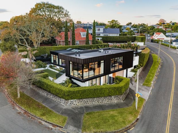 Aerial oblique view of a contemporary two-storey residential house on a landscaped corner site. The home has a strong rectangular form, dark vertical cladding, extensive floor-to-ceiling glazing, a low flat roof, and projecting upper-level volumes. Manicured hedges, stone retaining walls, paved paths, and established trees surround the property, with suburban streets and neighbouring homes visible beyond.