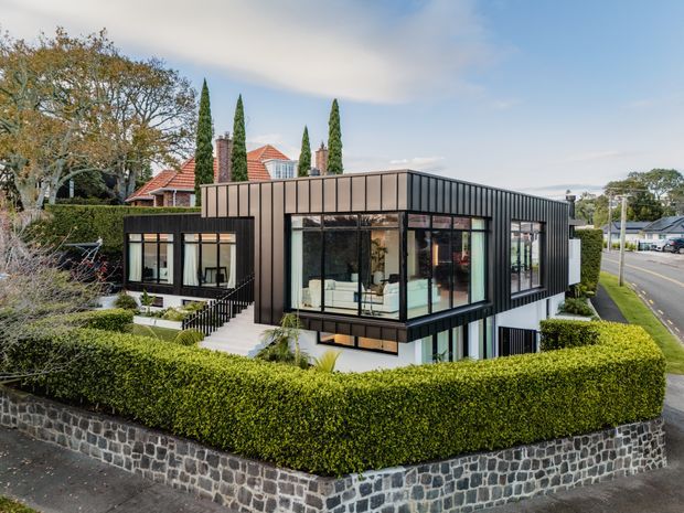 Exterior aerial view of a contemporary two-storey building with a dark standing-seam metal facade, extensive black-framed glazing, and a flat roof. The building sits behind a neatly clipped hedge and a low dark stone retaining wall, with a mature residential streetscape and a red-tiled traditional house visible behind it.