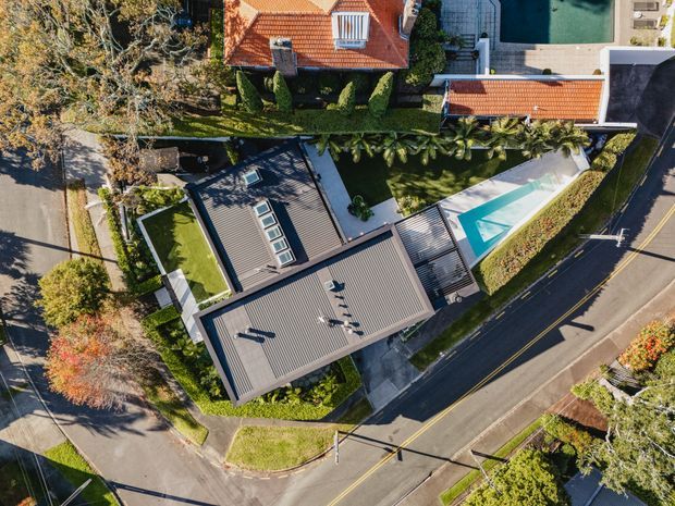 Aerial view of a contemporary residential property on a suburban corner site. The main dwelling has broad grey metal roofing with multiple skylights, adjoining roof volumes, landscaped gardens, paved paths, and a long turquoise swimming pool. Neighbouring homes, mature trees, streets, and road markings surround the property.
