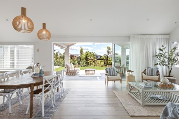 Bright, airy open-plan living and dining room with wide timber-look flooring, white walls, large windows and fully opening glazing to a covered outdoor area and garden. The dining area features a substantial timber table with white cross-back chairs, while the living zone has striped upholstered armchairs, a white coffee table, layered rugs and coastal pendant lights. Sheer curtains and abundant natural light create a relaxed coastal atmosphere.