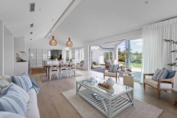 Bright, airy open-plan coastal-style living and dining area with a pale timber floor, white walls and ceiling, large glazed sliding doors, and sheer white curtains opening to a landscaped outdoor area. The living zone includes a pale upholstered sofa, striped blue-and-white cushions, timber-framed armchairs, a glass-topped coffee table, and a neutral rug. The adjacent dining area has a white table with cross-back chairs, while the kitchen is visible in the background. Pendant lights and abundant daylight create a relaxed, refined interior.
