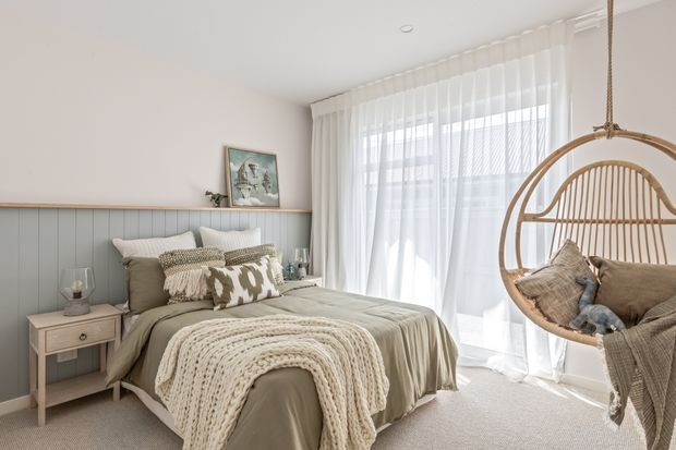 A bright, softly styled bedroom with a neatly made bed, pale sage-grey bedding, layered textured pillows, and a chunky knitted throw. The bed sits against a light blue-grey vertical panelled feature wall with a narrow picture ledge. A small light-wood bedside table, sheer floor-to-ceiling curtains, neutral carpet, and a suspended rattan hanging chair complete the relaxed coastal-inspired interior.