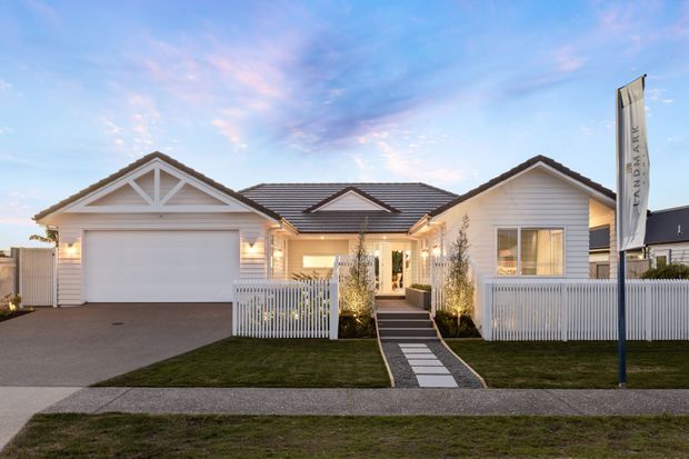 Street-facing exterior view of a single-storey suburban residence at dusk. The home has a symmetrical composition with a prominent front-facing gable over the double garage, a dark tiled hipped roof with smaller gabled dormer-like features, pale horizontal weatherboard cladding, white trim, a covered entry porch, white picket fencing, landscaped lawn, and a paved path leading from the footpath to the entrance. Exterior lighting creates a warm, welcoming appearance.