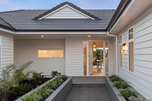 A contemporary residential entry courtyard viewed from outside, with light horizontal weatherboard cladding, a dark tiled gable roof, covered eaves, recessed ceiling lights, and a glazed front door opening into a warmly lit interior. Raised dark planter beds with tropical greenery line the gravel path to the entrance.