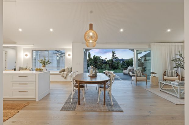 Bright open-plan coastal-style interior combining a white kitchen, dining area, and living room. The space features pale timber flooring, white cabinetry, large sliding glass doors opening to an outdoor deck, layered neutral furnishings, sheer curtains, and a woven pendant light over the dining table. Tropical landscaping is visible beyond the glazing.