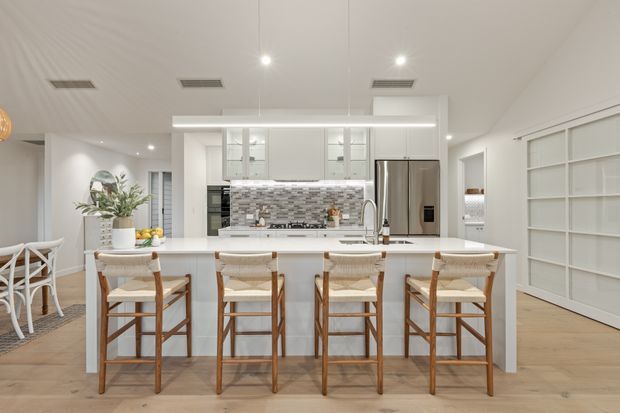 Bright, spacious open-plan kitchen centred on a large white island with four woven-seat timber bar stools. The room features white cabinetry, a patterned grey-and-white tiled splashback, stainless-steel appliances, pale timber flooring, integrated ceiling lighting and extensive built-in shelving. A dining area and hallway are visible in the background.