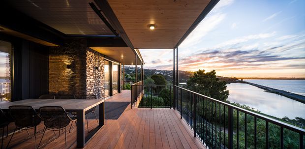 A contemporary covered balcony or terrace overlooking a calm waterfront at sunset. The space features warm timber decking and ceiling lining, black metal balustrades and framing, an outdoor dining setting, and a dramatic stone-clad wall beside large glazed openings. The terrace connects directly to a modern building and has elevated views across the water.