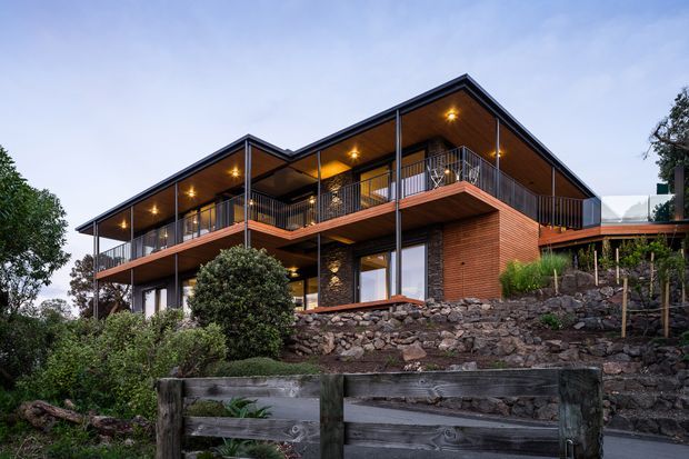 Exterior architectural photograph of a contemporary multi-level home set on a sloped, landscaped site. The building features dark metal framing, extensive covered balconies, warm timber soffits, exposed brick walls, large glazing, and stone retaining walls. The angular roof forms and illuminated verandas create a warm evening presentation.
