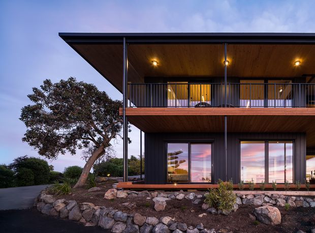Exterior dusk view of a contemporary two-storey house with dark vertical cladding, broad flat roof overhangs, timber-lined soffits and balcony, slim steel posts and balustrades, and large glazed doors. The home sits on a landscaped sloped site with a low natural-stone retaining edge and mature tree.
