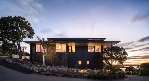 Twilight exterior view of a contemporary, low-profile house on a sloping site. The building has dark brick and vertical dark cladding, broad flat roof planes with deep overhangs, warm illuminated windows, and landscaped rock edging. A body of water and distant shoreline are visible beyond the property.